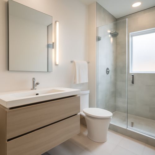 L'immagine mostra un bagno moderno e di design, con una combinazione di toni neutri e materiali eleganti. Il bagno &egrave; illuminato dalla luce naturale che entra attraverso una finestra, creando un'atmosfera luminosa e accogliente. Al centro, un mobile sospeso in legno chiaro con lavabo integrato, mentre a destra si trova una doccia a pavimento, separata da una parete in vetro trasparente. Un grande specchio senza cornice &egrave; posizionato sopra il mobile, mentre il pavimento &egrave; in piastrelle chiare, completando l'estetica minimalista e raffinata.