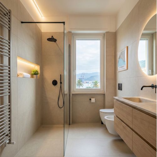 Fotografia di interior design che ritrae un bagno padronale moderno appena ristrutturato situato a La Spezia, Italia. L'ambiente &egrave; caratterizzato da grandi lastre di gres porcellanato effetto pietra color sabbia, un mobile lavabo sospeso in legno di rovere naturale e rubinetteria a contrasto in finitura nero opaco. Un'ampia doccia walk-in con vetro trasparente include una nicchia illuminata a LED. La finestra centrale offre una vista diurna sul porto e sul "Golfo dei Poeti", come confermato dalla mappa incorniciata sulla parete destra. L'illuminazione &egrave; un mix di luce naturale e strisce LED calde (3000K).