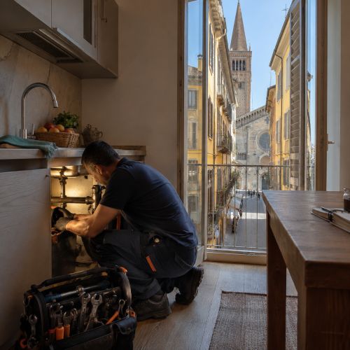Idraulico al lavoro in un appartamento del centro di Piacenza per una riparazione sotto il lavello con vista su una via storica.