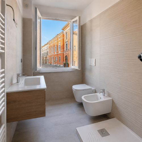 Bagno moderno in ristrutturazione a Forl&igrave; con sanitari nuovi, rivestimenti chiari, lavabo sospeso e finestra aperta su una via del centro storico.