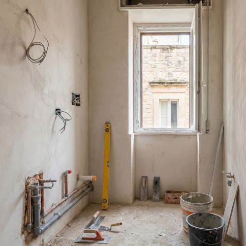 Un bagno in fase di cantiere, con pareti in intonaco grezzo e i nuovi impianti idraulici ed elettrici a vista. Attrezzi da lavoro e secchi sono sparsi sul pavimento. La finestra inquadra un tipico edificio storico di Bari, localizzando la scena.